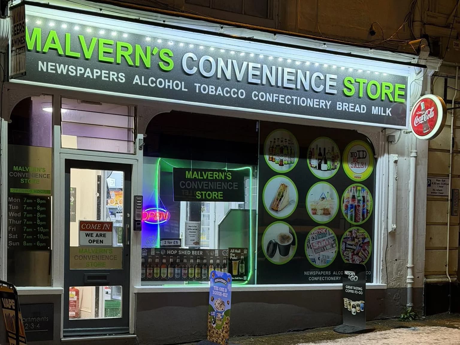 The front of Malvern’s Convenience Store at night, its green and white sign lit above the door and a window of drinks, sandwiches, newspapers and sweets